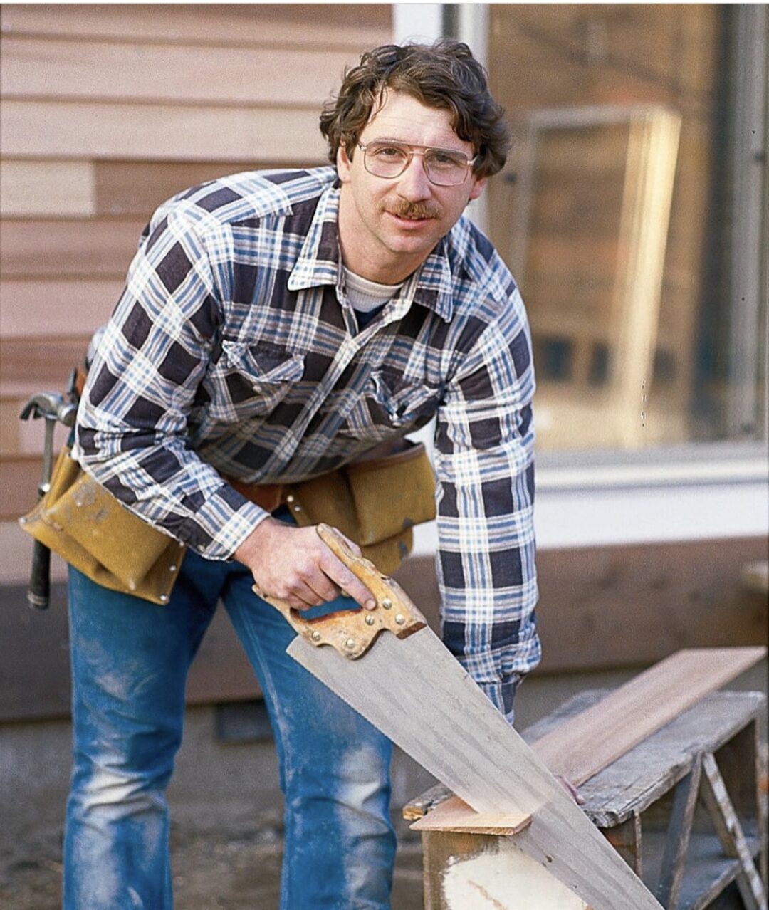 A man wearing glasses, a plaid shirt, and a tool belt is sawing a piece of wood outdoors. He is leaning over the wood and looking at the camera, with a house wall in the background.