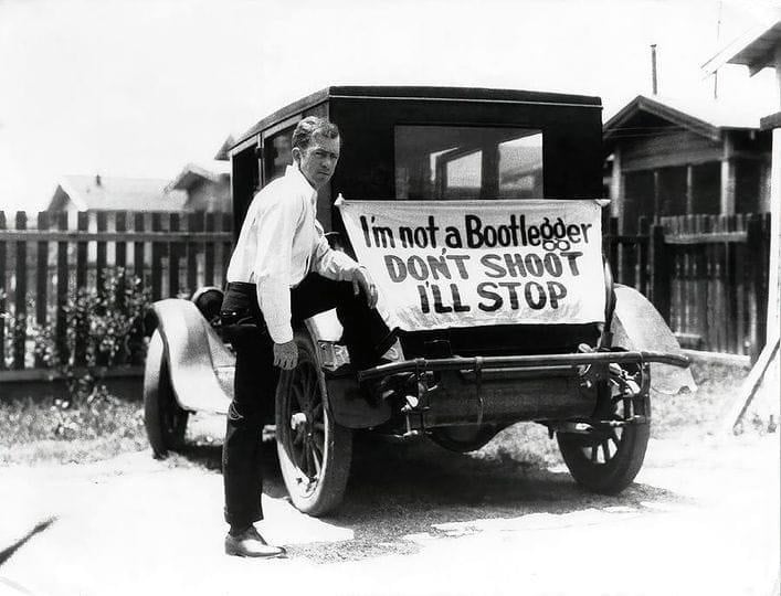 A man stands beside an old car with a sign on the back that reads, "I'm not a Bootlegger. Don't shoot. I'll stop." The scene appears to be from the early 20th century, in a residential neighborhood.