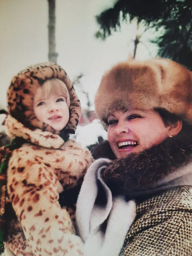 A woman in a fur hat and scarf smiles while holding a child dressed in a leopard print winter outfit. Snow and palm trees are visible in the blurred background.