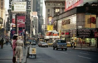 A busy city street scene in Times Square, New York, with people walking, cars and yellow taxis driving, and bright advertisements and billboards for products and movies covering the buildings.