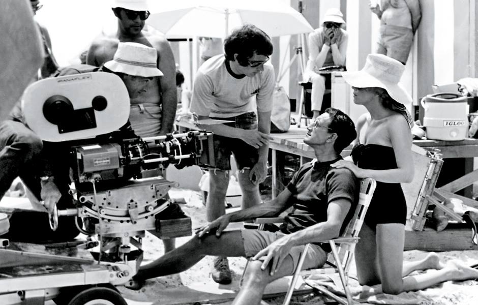 A black-and-white photo of a film crew on a beach set. Several people gather around a large movie camera, with one man seated and a woman in a swimsuit and sunhat standing beside him, as others observe or adjust equipment.