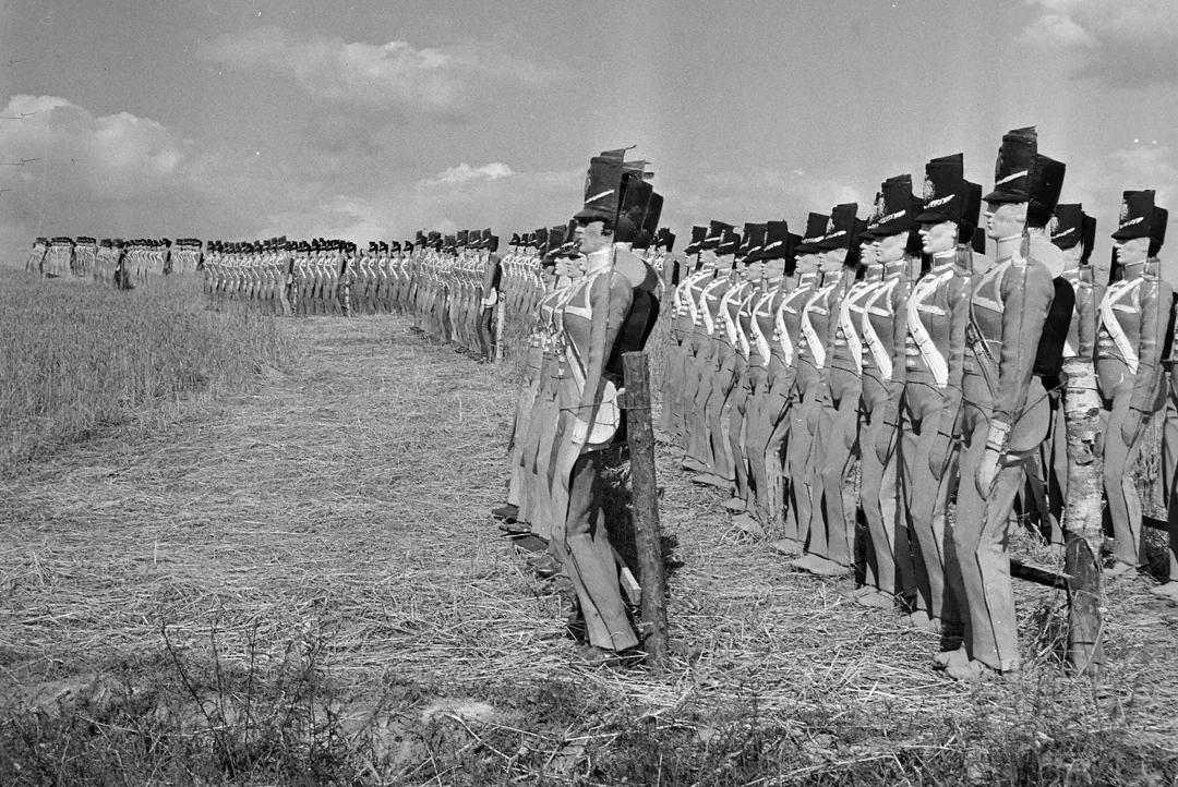 Rows of life-sized scarecrows dressed in military uniforms and hats stand in a field, arranged to resemble marching soldiers under a partly cloudy sky.