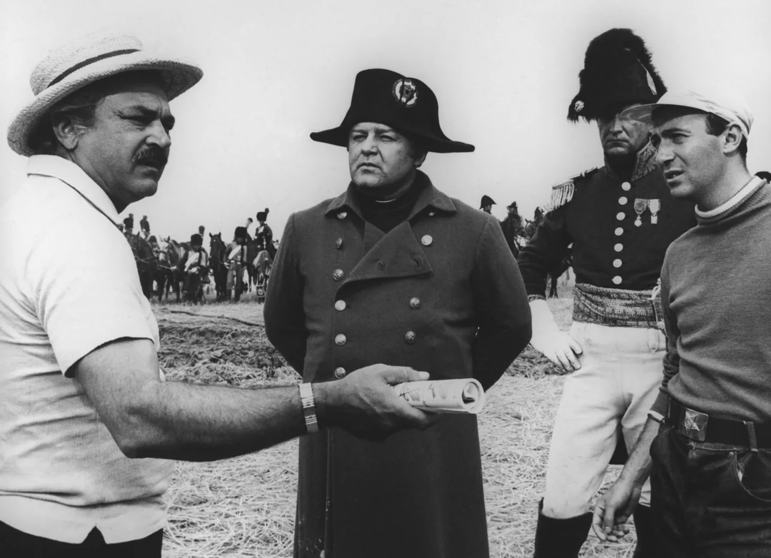 A black-and-white photo shows four men on a film set. One man wears a straw hat and holds a film canister, while two others are dressed in historical military uniforms, and a fourth man wears casual clothes. Extras are visible in the background.