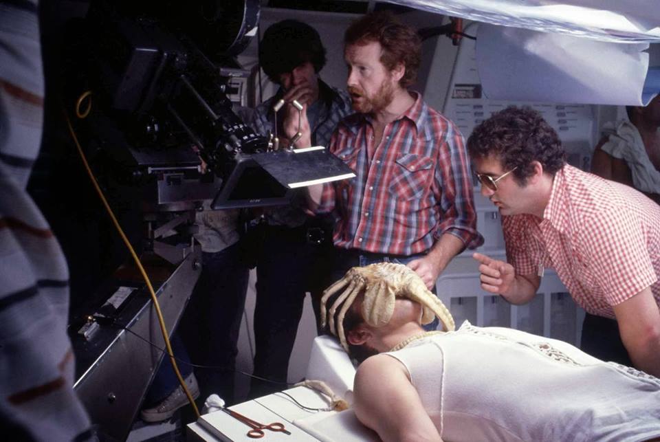 Three men, including a cameraman and two crew members, are filming a scene with an actor lying on a table with a science fiction creature attached to their face. The setting appears to be a movie set.