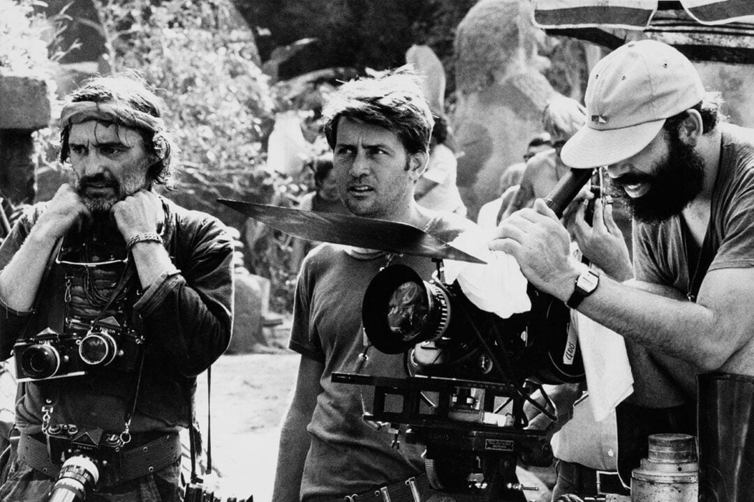 Three men work on a film set outdoors. One adjusts his headset with cameras around his neck, another stands in the middle looking focused, and the third operates a film camera, cleaning the lens, all surrounded by filming equipment.