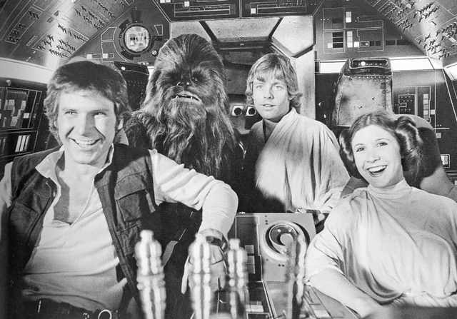 Four "Star Wars" characters—Han Solo, Chewbacca, Luke Skywalker, and Princess Leia—smile inside the Millennium Falcon’s cockpit, surrounded by sci-fi controls in a black-and-white photo.