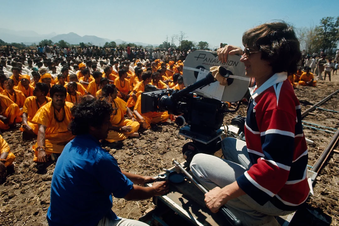 A film crew shoots a scene outdoors; a camera operator films rows of people dressed in orange, seated on the ground under clear blue skies, while another crew member adjusts equipment beside them.