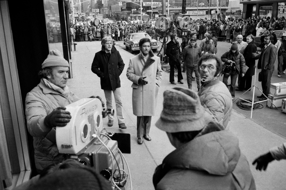 A film crew shoots a scene on a busy city street, with camera operators in the foreground, an actor in a long coat at the center, and a large crowd of onlookers behind barriers in the background.