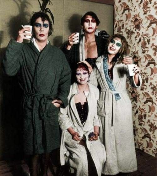 Four people in bathrobes and dramatic makeup pose together indoors, holding cups and smiling. The background features floral wallpaper and a plant. Their expressions and attire suggest a playful, theatrical atmosphere.