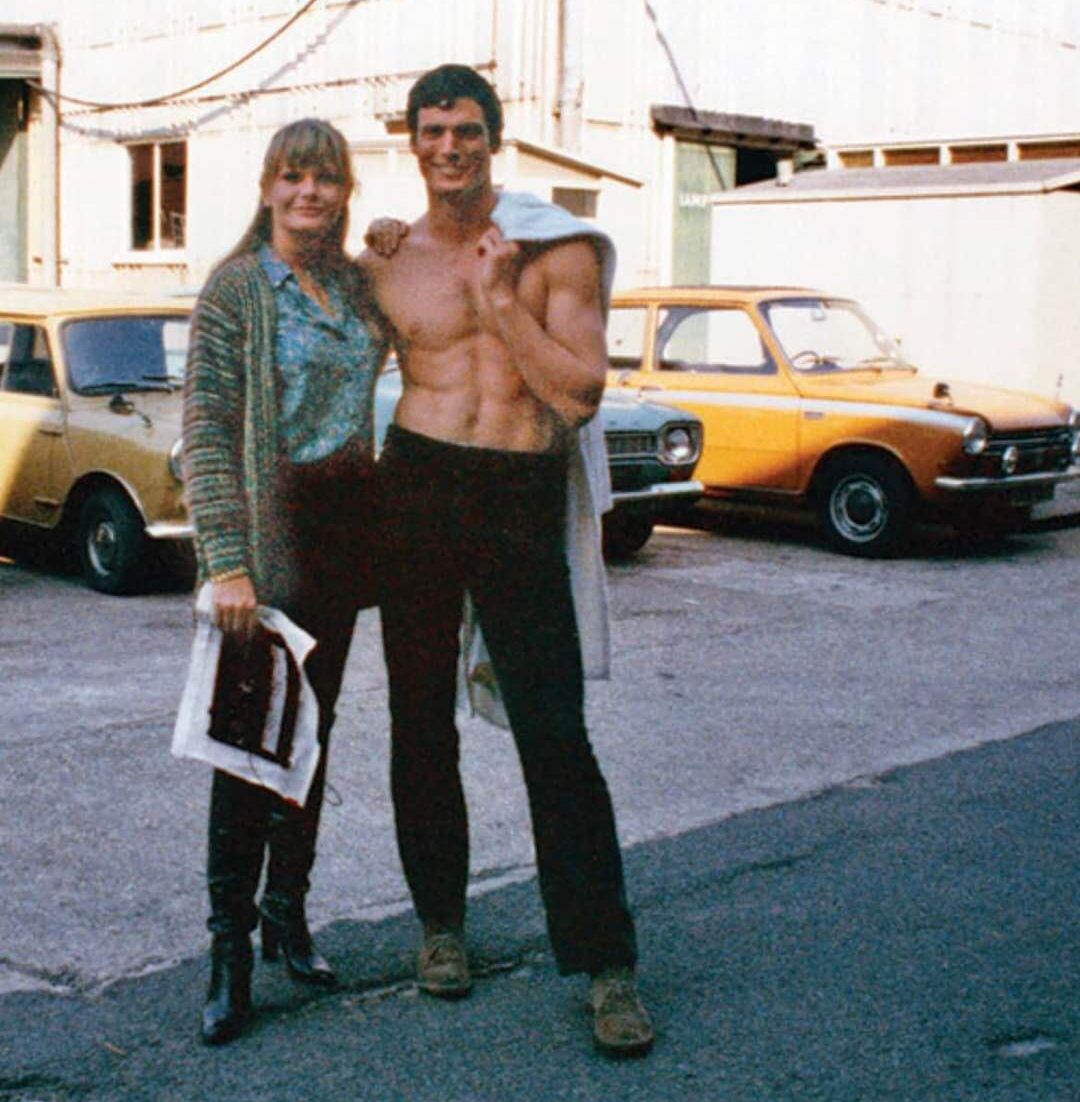 A woman stands next to a smiling shirtless man with a jacket over his shoulder in a parking lot, with vintage cars and an industrial building in the background.