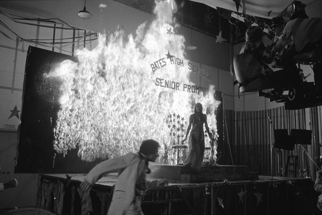 A woman in a dress stands on a stage engulfed in flames at a high school prom, while a man runs away. The backdrop reads “Bates High School Senior Prom.” Film crew and camera equipment are visible nearby.