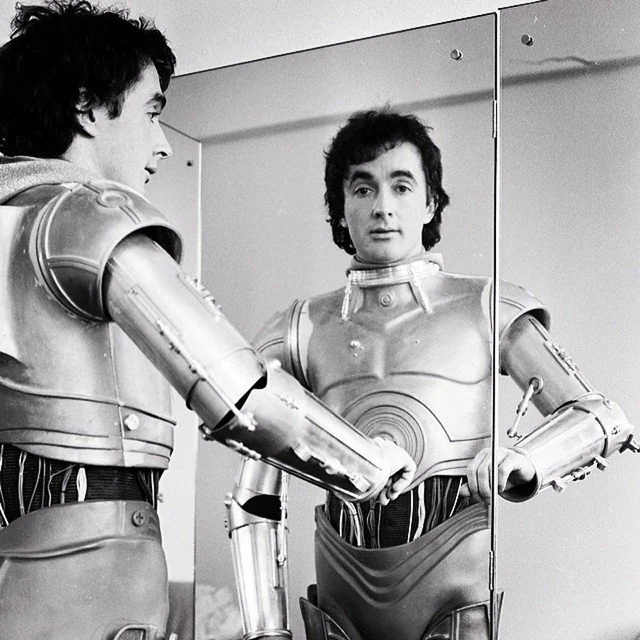 A man in a robot costume stands in front of a mirror, looking at his reflection. The costume has metallic arms and chest plates, and the mirror shows both his face and the details of the suit.