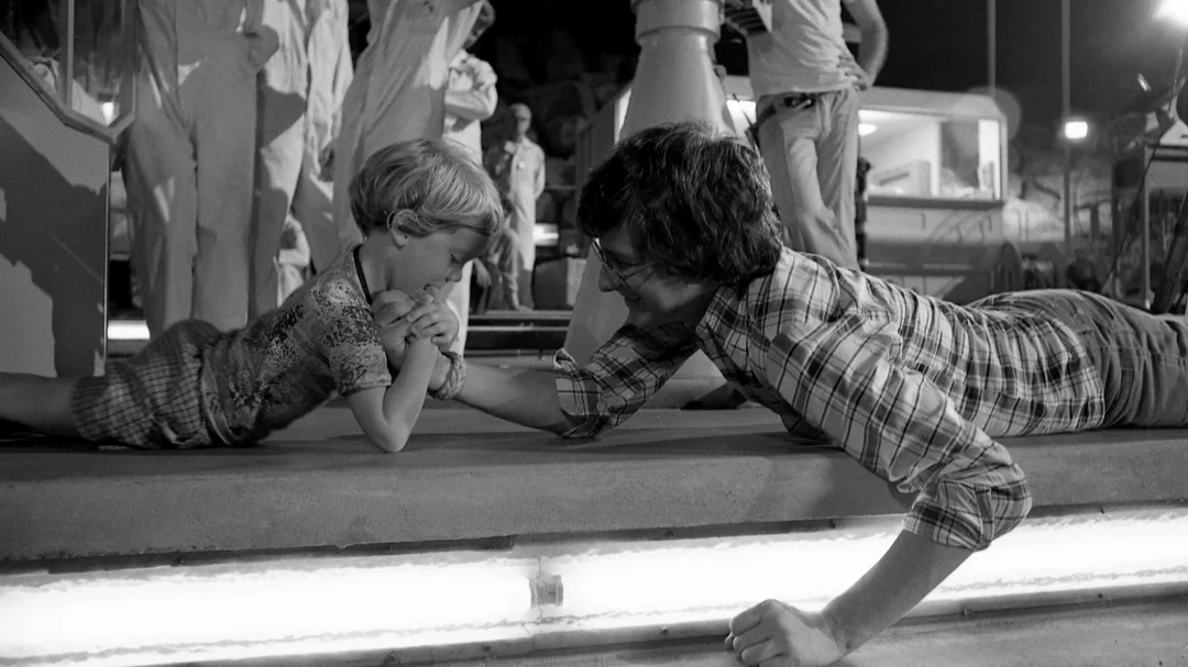A young boy and an adult man arm wrestle playfully on the ground, smiling at each other, while people stand and watch in the background. The image is in black and white.
