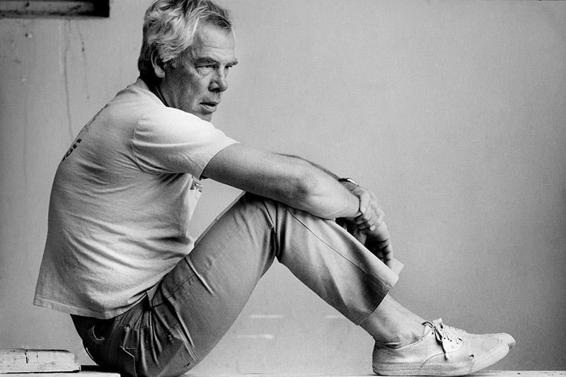 A man with gray hair sits sideways on the floor against a light background, wearing a light t-shirt, pants, and sneakers, looking thoughtfully into the distance.