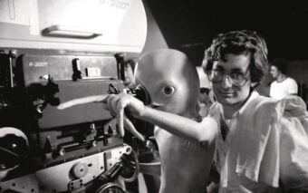 A man with glasses stands next to an alien puppet, both looking at and pointing towards a film camera on a movie set, surrounded by crew members in the background.