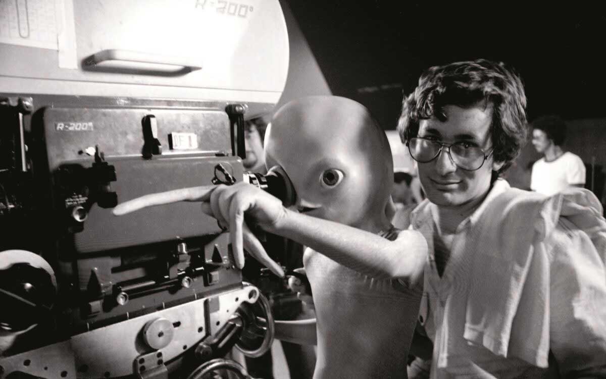 A man with glasses stands next to an alien puppet, both looking at and pointing towards a film camera on a movie set, surrounded by crew members in the background.