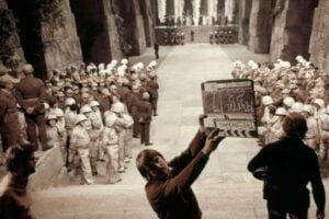 Behind the scenes of a film set resembling a grand stone hall, with actors in white armor and military uniforms standing in rows. A crew member holds a clapperboard in the foreground.