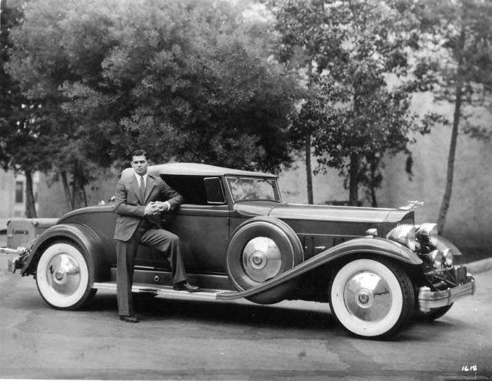A man in a suit leans against a vintage luxury car with large whitewall tires, parked on a street with trees in the background. The image is black and white, evoking a classic, early-20th-century style.