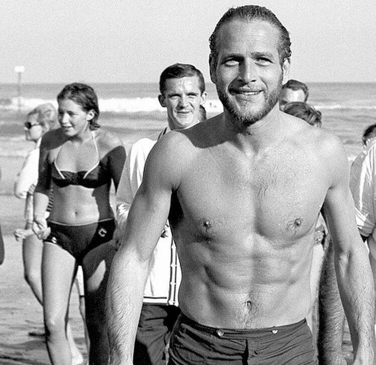 A smiling, fit man in swim trunks walks on a beach, with people in swimsuits and casual clothes behind him. The ocean and waves are visible in the background. The image is in black and white.