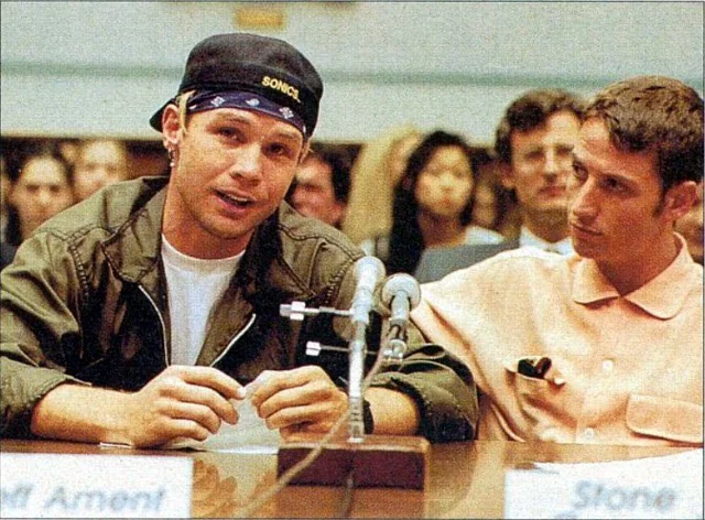 Two young men sit at a table with microphones, speaking at a public hearing. One wears a cap and jacket, gesturing as he talks; the other wears a light shirt and listens attentively. Blurred audience is visible in the background.