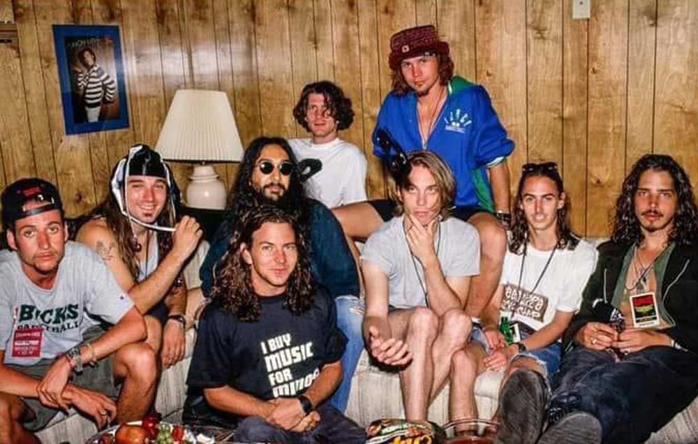 Nine men with long hair sit and stand closely together in a wood-paneled room, some smiling at the camera. They are casually dressed, with a lamp, framed poster, and snacks on the table in front of them.