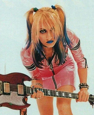 A person with blonde and blue hair in pigtails, wearing bold blue lipstick and a shiny pink outfit, leans forward holding an electric guitar. The background is white.