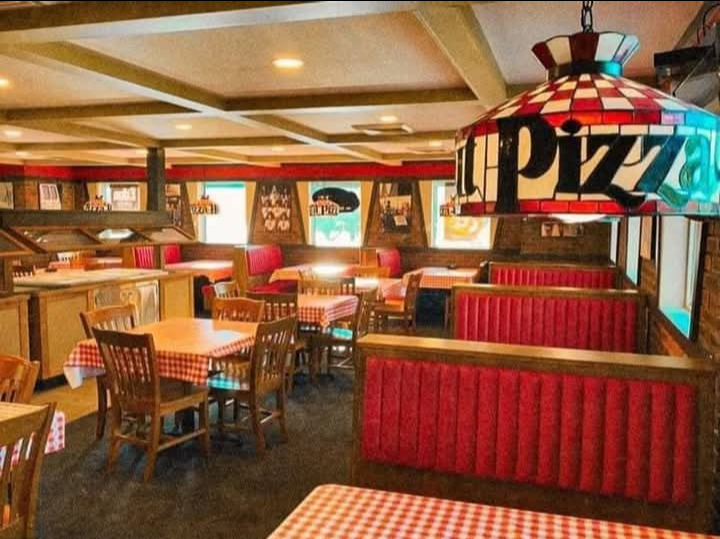 A cozy pizzeria with red booth seating, wooden chairs, checkered tablecloths, and a colorful stained-glass lamp that says “pizza.” The decor includes vintage signs and large windows.