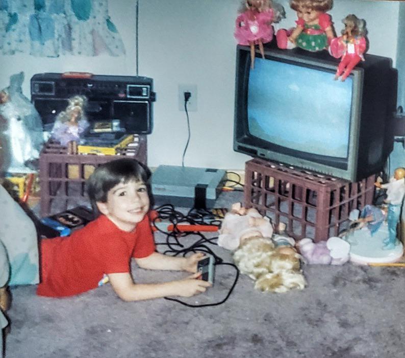 A child in a red shirt lies on the carpet playing video games on a small TV, surrounded by dolls and toys. The TV sits on a crate with stuffed dolls on top. Nearby, there’s a stereo system and more toys in the background.