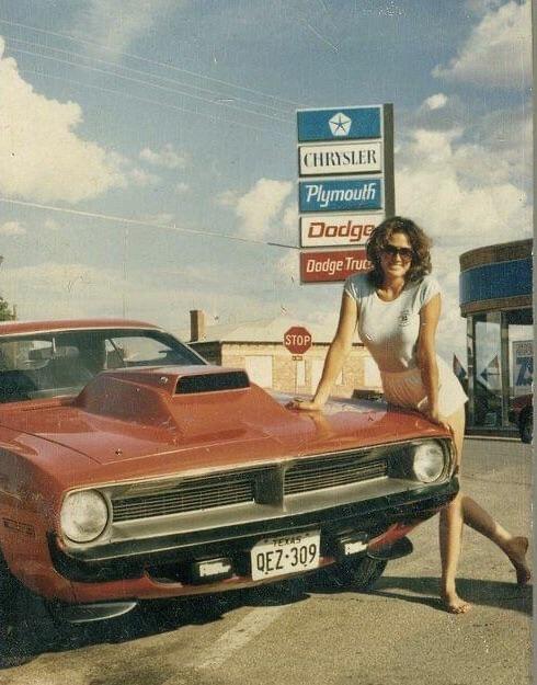 A woman in a white dress leans on the hood of a red classic car parked near a Chrysler, Plymouth, Dodge, and Dodge Trucks dealership under a blue sky.