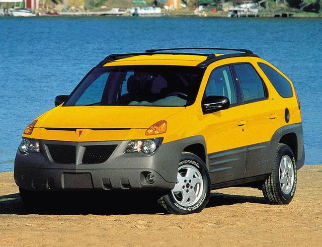 A yellow and gray Pontiac Aztek SUV is parked on sandy ground by a body of water, with trees and boats visible in the background.