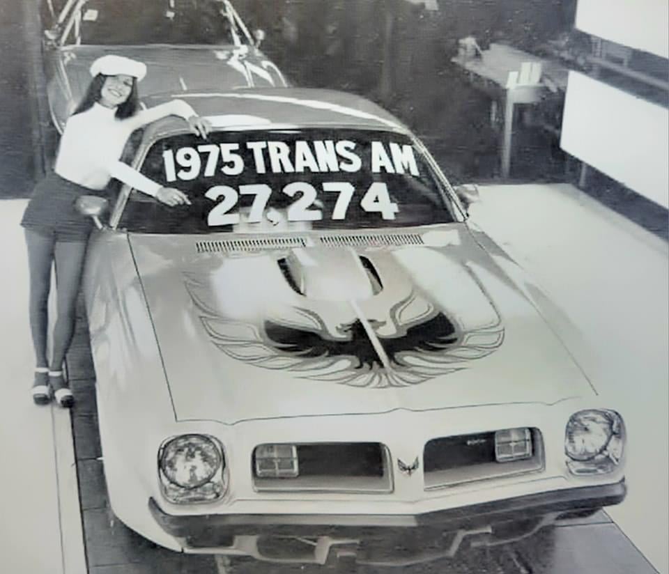 A woman in a hat and shorts leans on a 1975 Trans Am car with a large firebird decal on the hood and a sign reading "1975 TRANS AM 27,274" on the windshield. The photo is black and white.