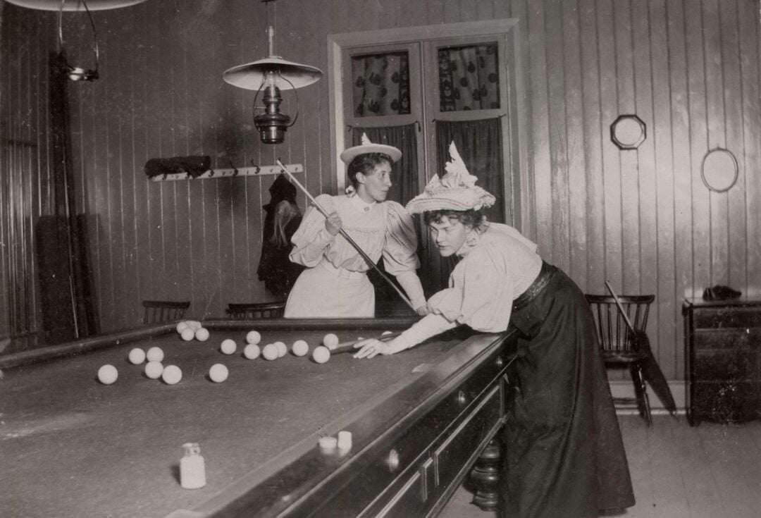 Two women in vintage clothing play billiards in a wood-paneled room. One lines up a shot with a cue stick while the other leans over the table. The room has chairs, mirrors, and overhead lighting.