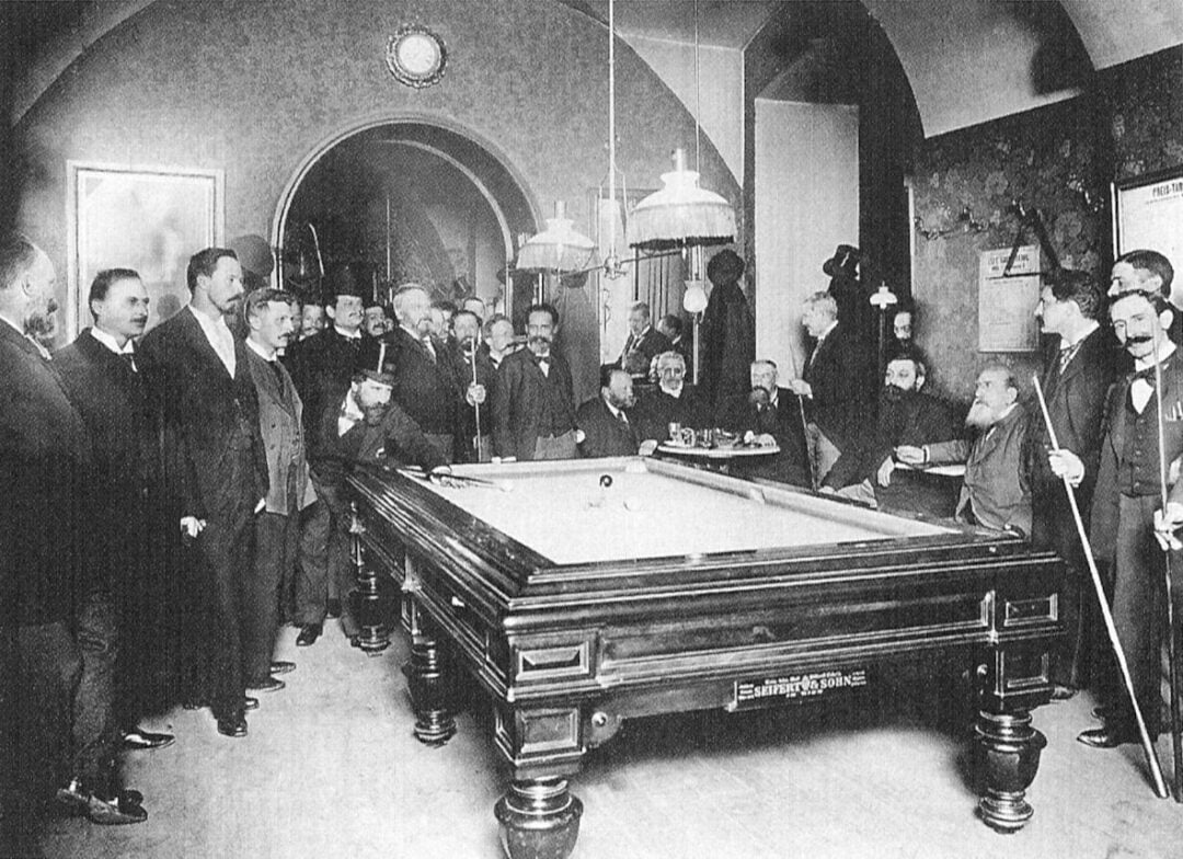 A black and white vintage photo showing a group of men in formal attire gathered around a billiard table, watching a game in an ornate room with arched ceilings and hanging lights.