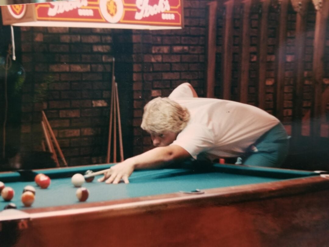 A person with blond hair and a white shirt leans over a pool table, aiming a cue stick at the balls. Pool cues rest against a brick wall in the background, and a beer sign hangs above.