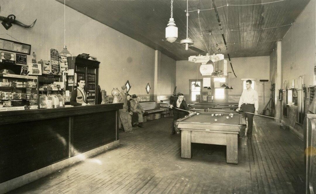 A black-and-white photo of an old-fashioned bar or general store with a billiard table. Two men play pool while a woman stands behind the counter and two people sit on a bench in the background. The room has high ceilings and wood floors.