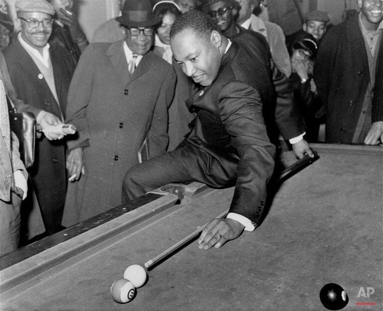 A man in a suit leans over a pool table to take a shot, surrounded by a group of people watching him and smiling. The scene appears lively and social.