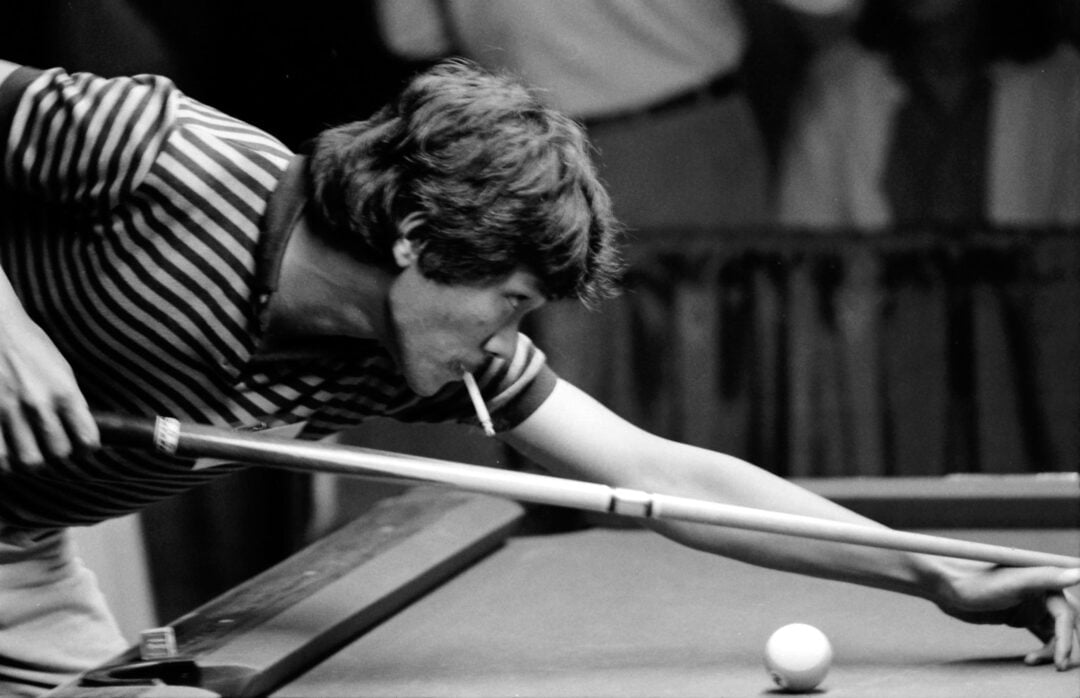 A man in a striped shirt leans over a pool table, aiming his cue at the cue ball while holding a cigarette in his mouth.