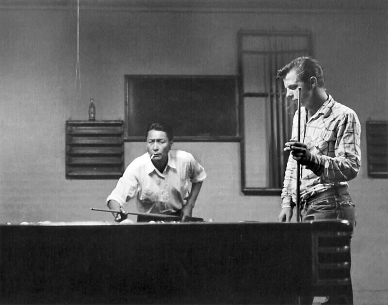 Two men stand by a billiards table indoors. One man is taking a shot, focused on the game, while the other watches, holding a cue stick. The room is dimly lit, with windows and a bottle on a ledge in the background.