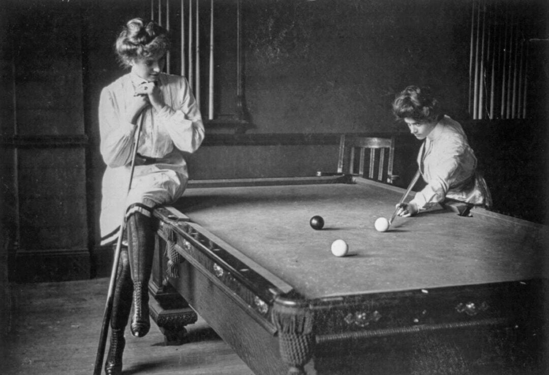 Two women in vintage clothing play billiards; one leans on a cue stick watching while the other lines up a shot at a pool table. Three balls are visible on the table; cues rest on the wall in the background.