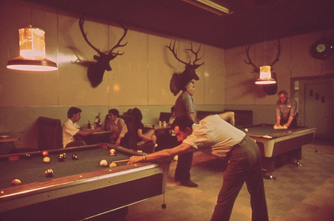 A man lines up a shot on a pool table in a dimly lit bar with two pool tables, people seated at booths, and deer heads mounted on the wall. Two men play pool while others watch and socialize in the background.