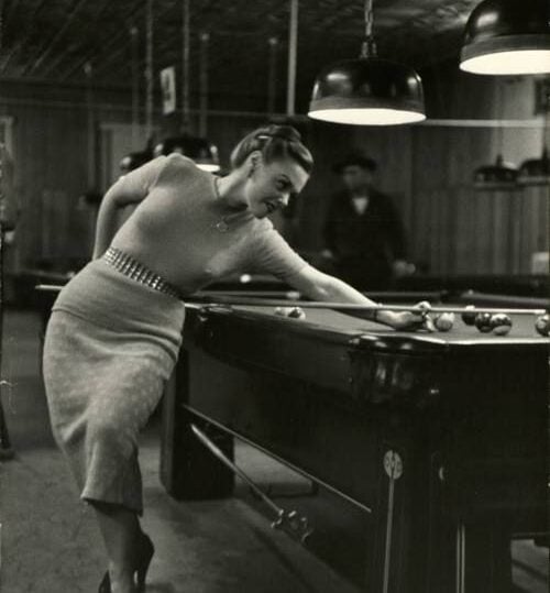 A woman in a fitted dress leans over a pool table to take a shot, concentrating on her aim. Other people and pool tables are visible in the background under hanging lamps. The photo is in black and white.