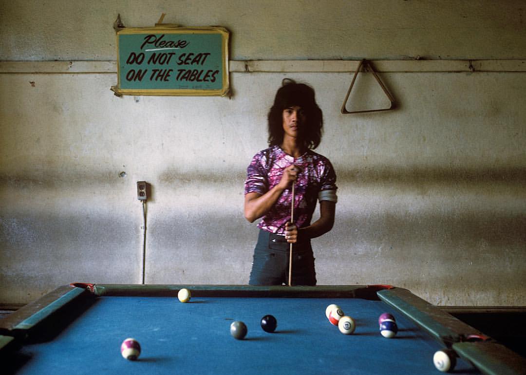 A person with long hair, wearing a purple patterned shirt, stands behind a pool table holding a cue stick. A sign on the wall reads "Please DO NOT SEAT ON THE TABLES." Several balls are scattered on the table.