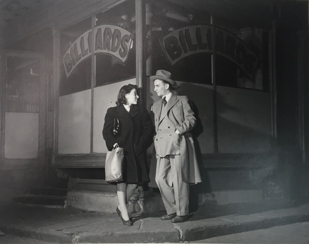 A woman and a man in mid-20th-century clothing stand and smile at each other outside a storefront with large signs reading “BILLIARDS.” It is nighttime, and the scene is lit dramatically with deep shadows.