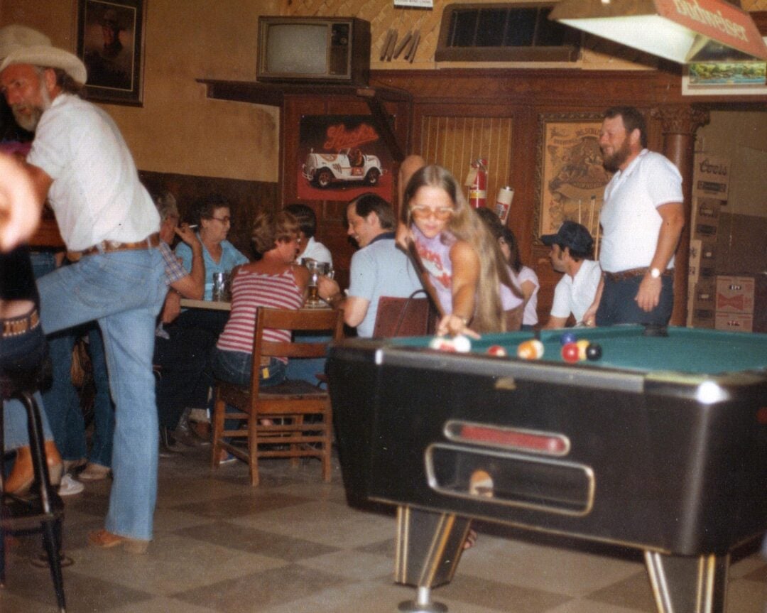 A girl in sunglasses prepares to shoot pool in a busy, vintage bar with people chatting at tables and a man in a cowboy hat sitting at the counter. The scene has a retro, casual atmosphere.
