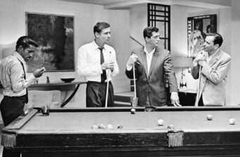 Four men in suits stand around a pool table, holding cues and talking. One man lights a cigarette while the others watch and chat in a stylish, mid-century modern room with artwork on the walls.