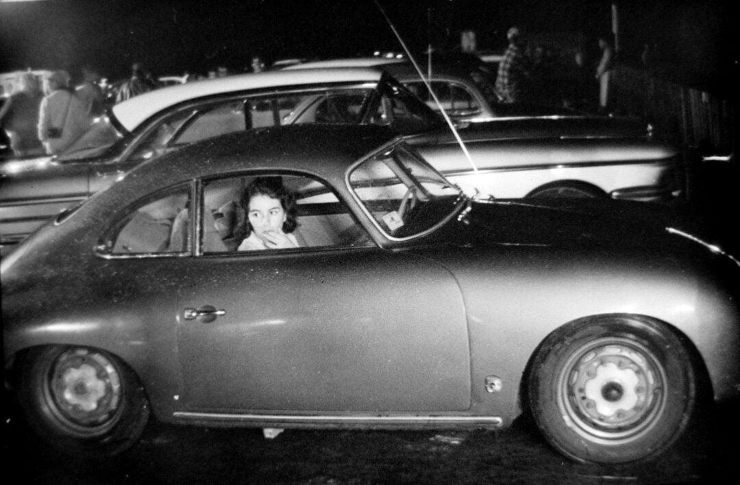 A black-and-white photo showing a woman sitting in the driver's seat of a vintage car at night, looking out the window. Other classic cars and people are visible in the background.