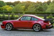 A classic red Porsche sports car is parked on a gravel driveway with green trees, bushes, and a wooden fence in the background at sunset.