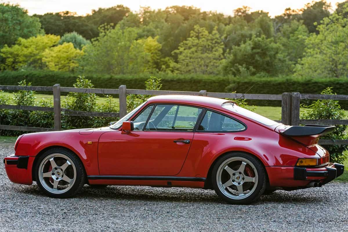 A classic red Porsche sports car is parked on a gravel driveway with green trees, bushes, and a wooden fence in the background at sunset.
