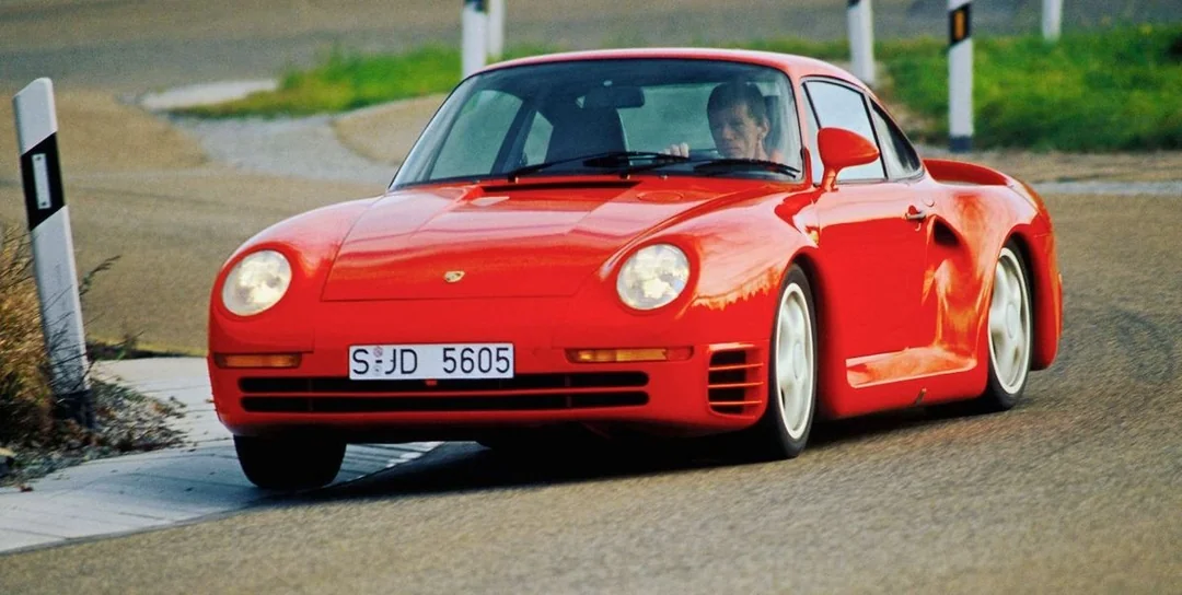 A red Porsche 959 sports car with a white license plate is driving around a curve on a paved road, surrounded by green grass and roadside markers.