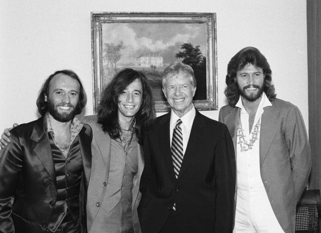Four men stand side by side, smiling, in front of a framed painting. Three wear stylish suits with open collars, and one wears a striped tie and suit. The photo appears to be black and white and from the 1970s.